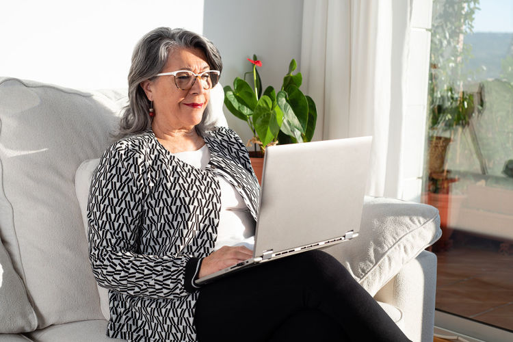 Content respectable mature businesswoman wearing trendy elegant clothes and eyeglasses browsing modern netbook in light office