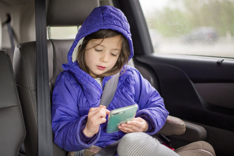 A small child wits in her carseat on a trip playing with a cell phone