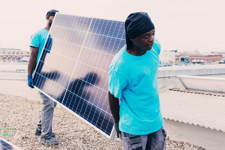 African american workers walking with solar panel on sunny day in industrial area