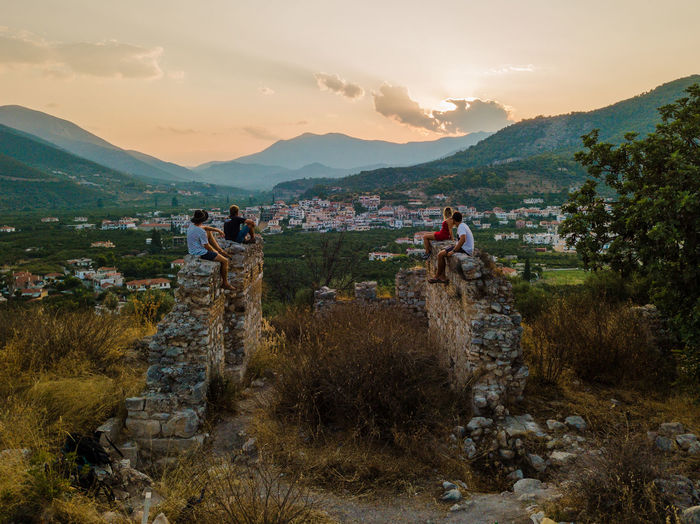Friends sitting on old ruins against sky at sunset