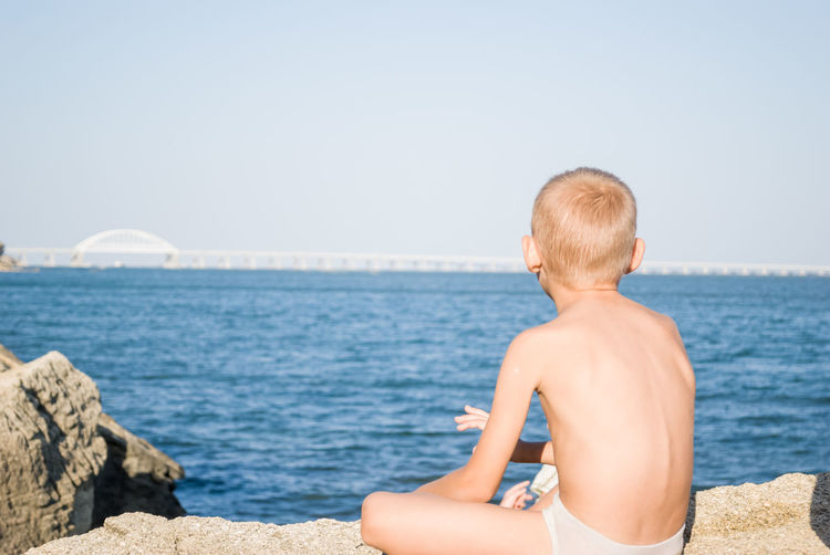 Rear view of boy sitting on rock by sea against sky