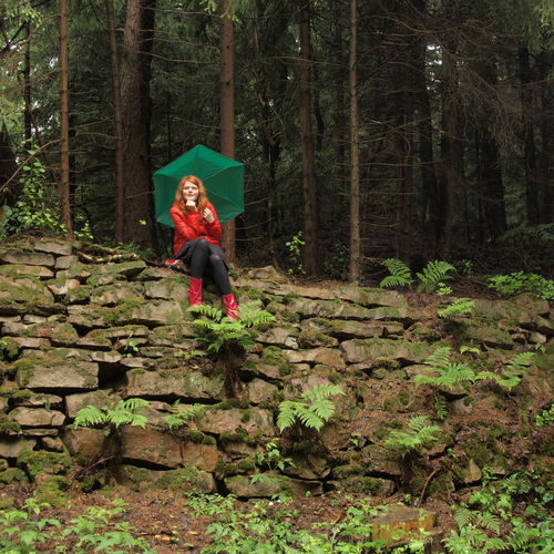 Young woman in raincoat with umbrella sitting on stone wall at forest