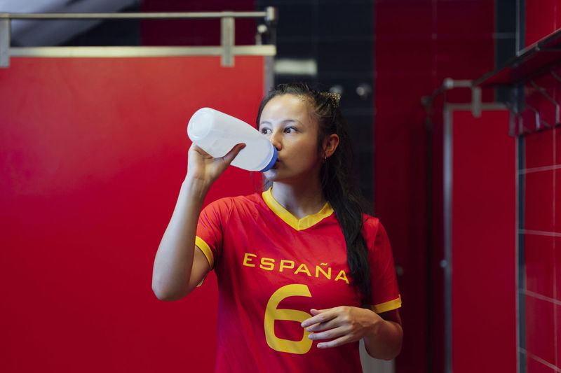 Young woman drinking water while standing against yellow wall