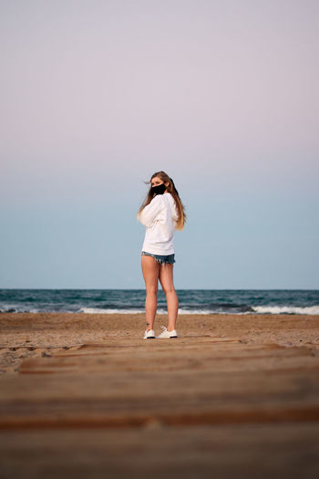 Back view of female tourist wearing protective mask standing on sandy beach near sea against sunset sky and looking over shoulder during coronavirus epidemic