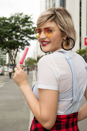Portrait of smiling woman holding ice cream while standing on footpath in city