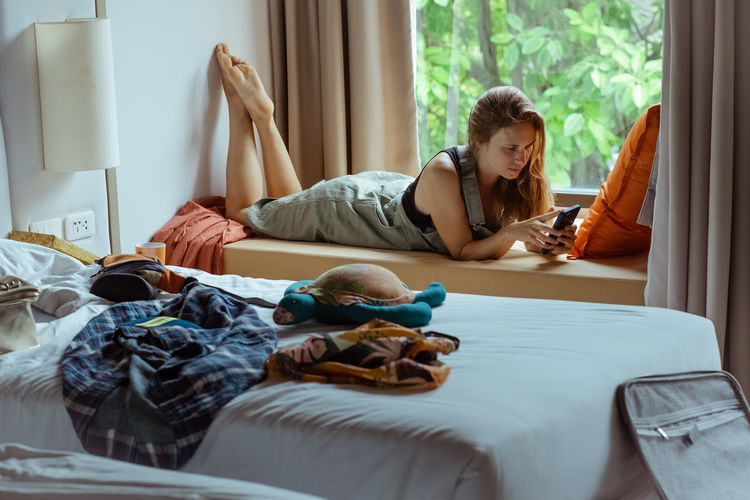 Girl student lying by the window and talking on mobile phone in room with mess.