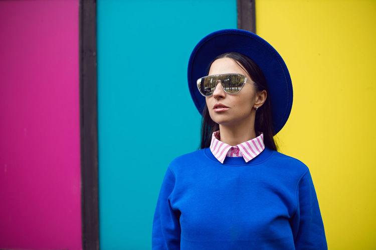 Fashionable brunette woman in a blue warm hat and sweater and sunglasses stands against background