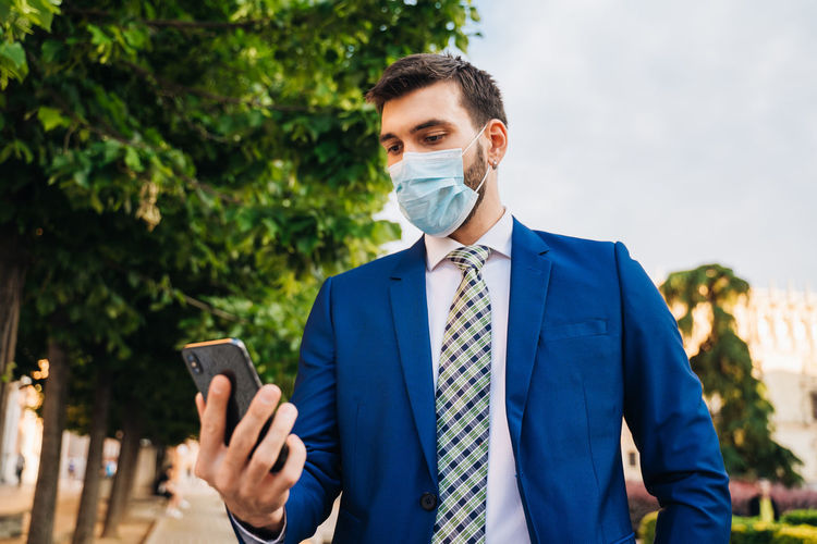 Young elegant businessman with a surgical mask looks at the phone in the park