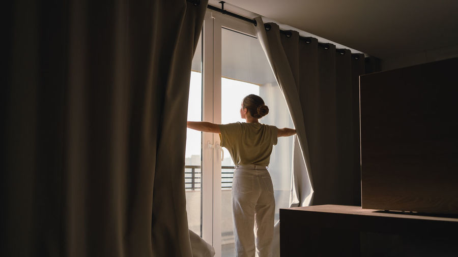 Back view of unrecognizable young female in casual clothes opening blackout curtains and admiring modern city during holidays in hotel