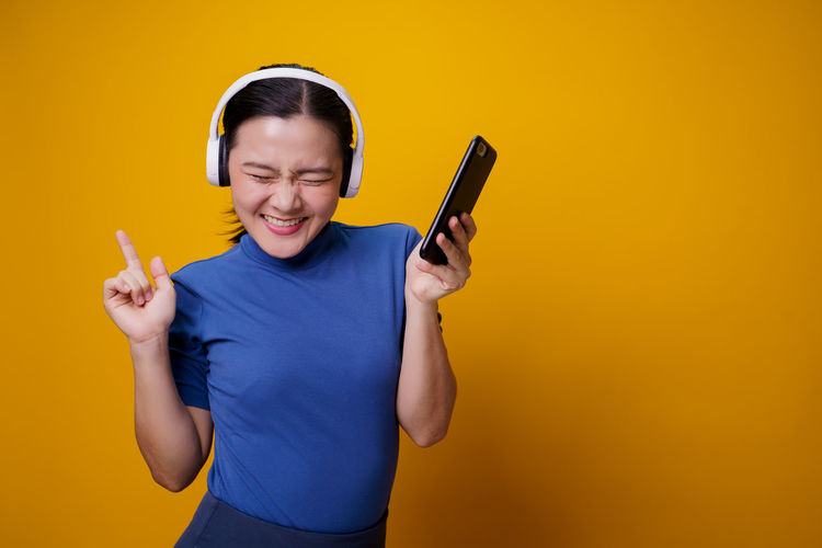 Young woman using phone while standing against yellow background