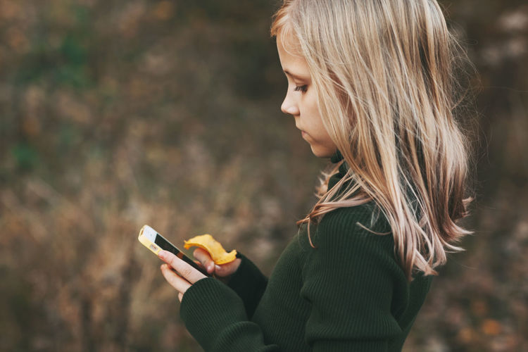 Blonde teenage girl stands with the phone and writes a message in the park outdoors.
