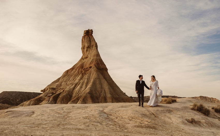 Happy groom and bride walking holding hands near mountain against cloudy sundown sky in bardenas reales natural park in navarra, spain