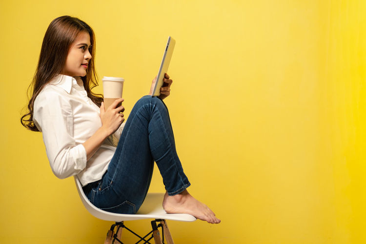 Young woman using mobile phone while sitting on chair against yellow wall