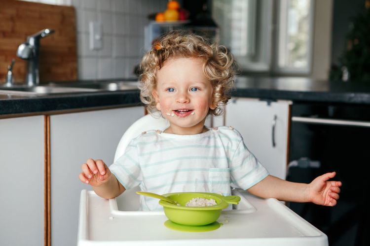 Caucasian curly kid boy sitting in high chair eating cereal puree with spoon. healthy eating 