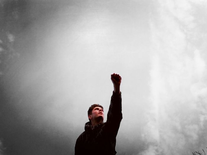 Low angle view of young man with hand raised standing against cloudy sky