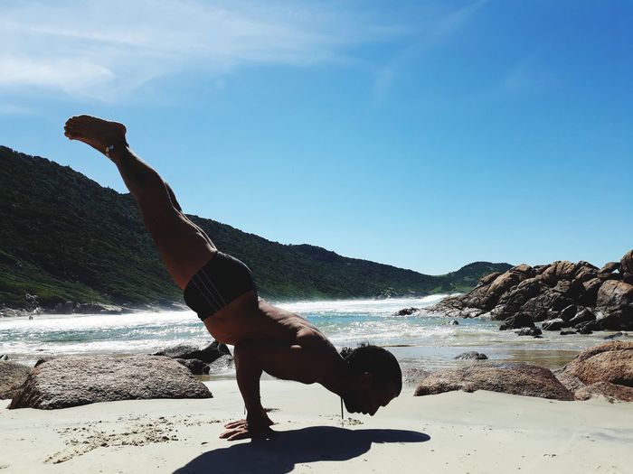 Full length of shirtless man practicing handstand at beach against blue sky