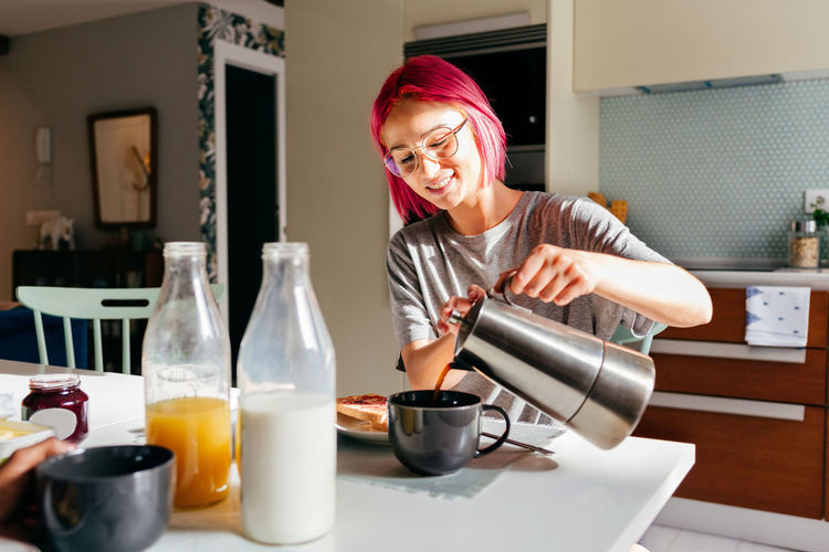 Young woman with dyed hair smiling and filling mug with fresh coffee while having breakfast in morning at home