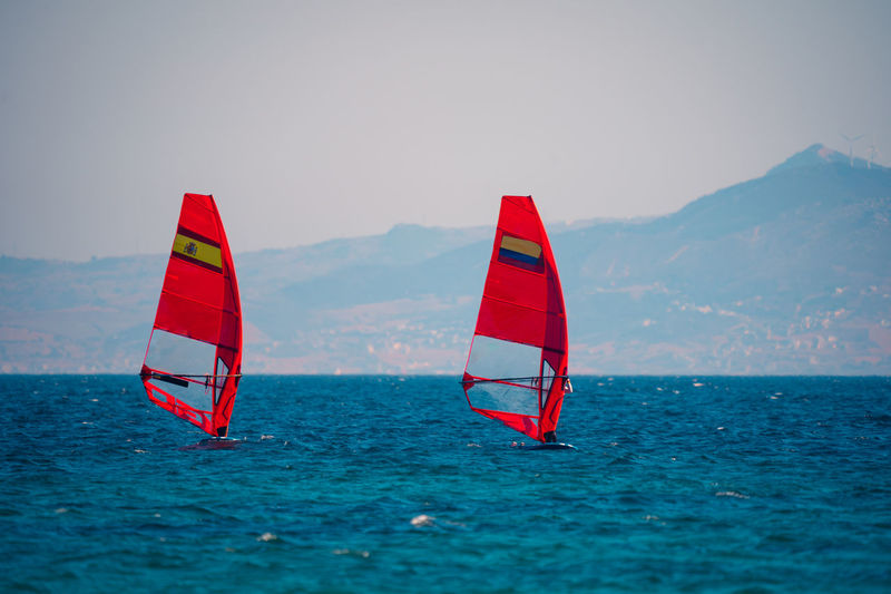 Red sails of windsurfers on rippling blue water of sea bay with blurred mountains on background in sunlight