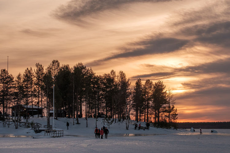 People walking on snow covered field against sky during sunset