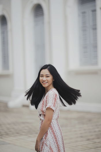 Portrait of smiling young woman tossing her hair while standing against building