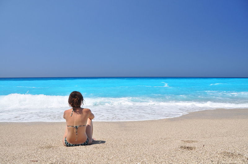 Rear view of woman in bikini sitting at sandy beach against clear sky