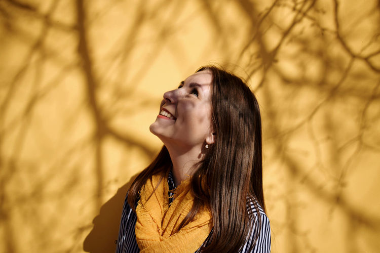 Smiling young woman looking up while standing against yellow wall