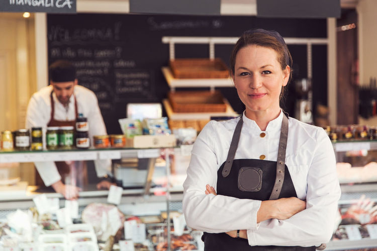 Portrait of confident owner standing arms crossed while colleague working in background at grocery store