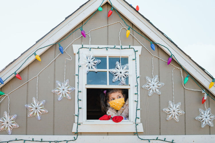 Young girl in face mask looking out window christmas decorations