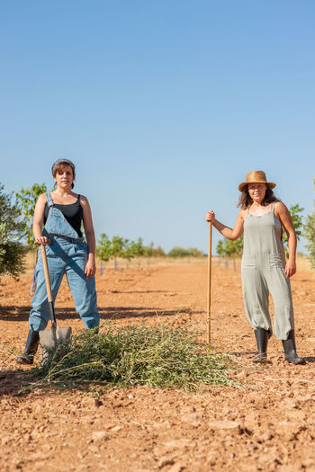 Women in overalls and rubber boots using rake for harvesting dried grass while having fun in village in summer