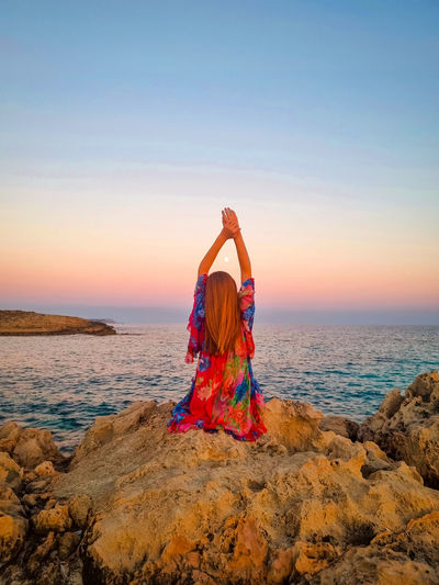 Rear view of woman doing yoga at beach against sky during sunset