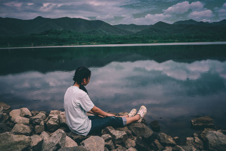 Man sitting on rock by lake against sky