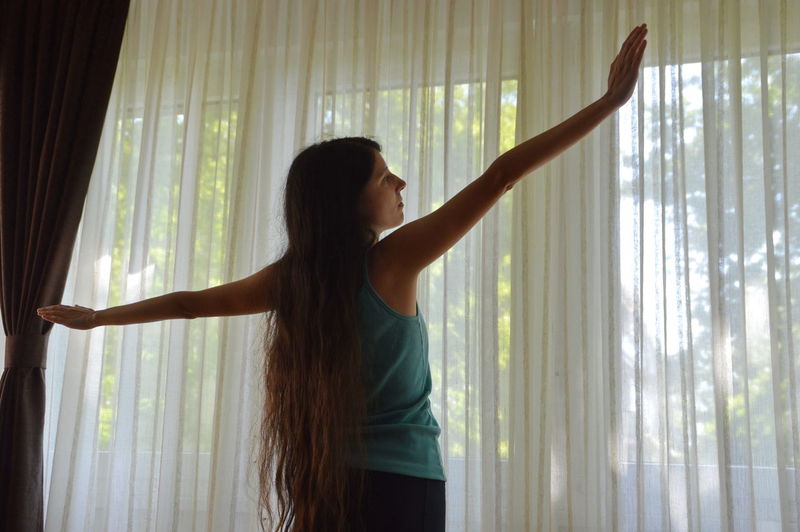 Rear view of woman with arms outstretched standing against curtain at home