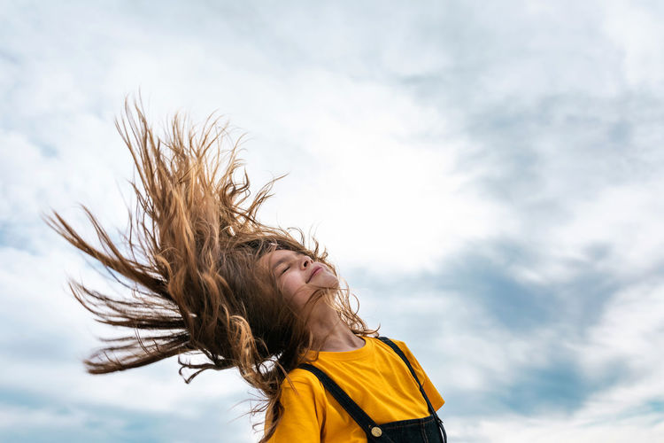 Side view from below of peaceful teenager throwing long hair on background of cloudy sky in summer