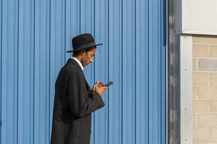 Young man in hat using mobile phone while standing by corrugated iron on sunny day
