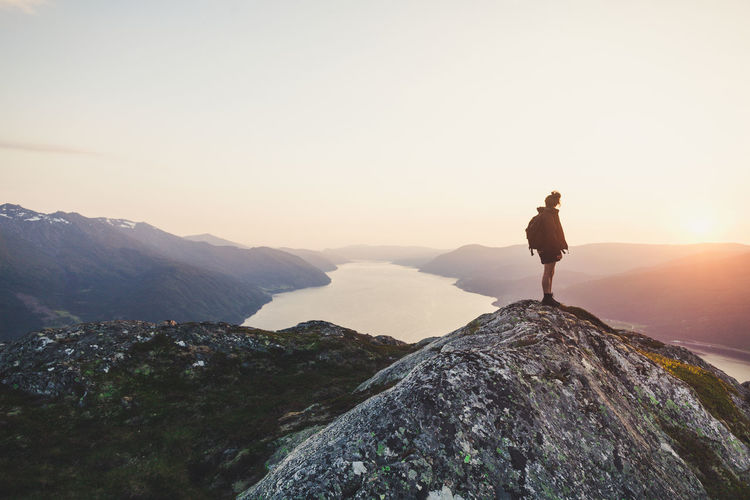 Rear view of woman looking at river amidst mountains against sky during sunset