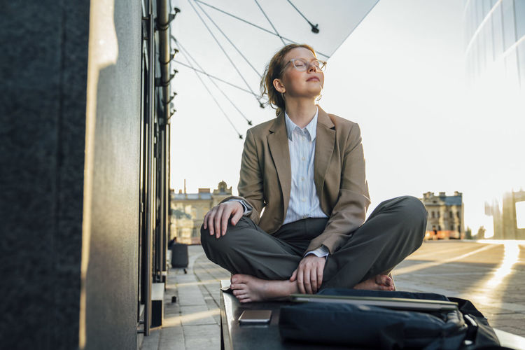Young businesswoman with eyes closed sitting cross-legged by office building