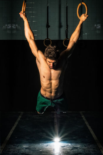 Full length strong shirtless guy doing exercise on gymnastic rings during intense workout in dark gym