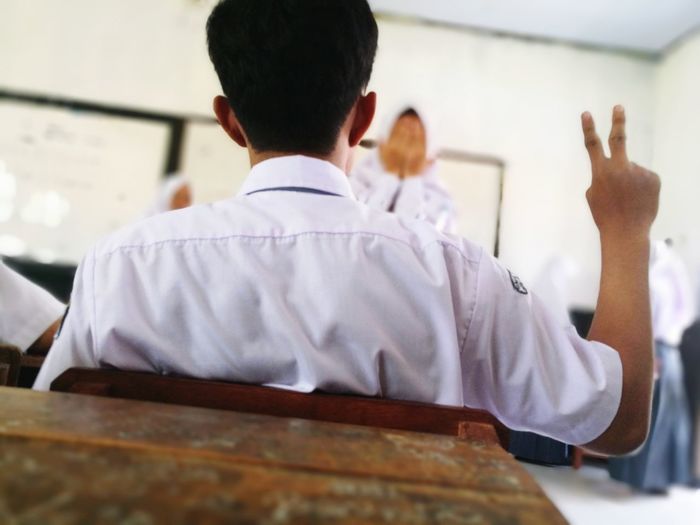 Rear view of teenage boy gesturing while siting on bench in classroom