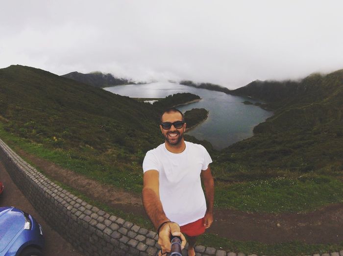 Portrait of happy man taking selfie while standing on retaining wall against lagoa do fogo
