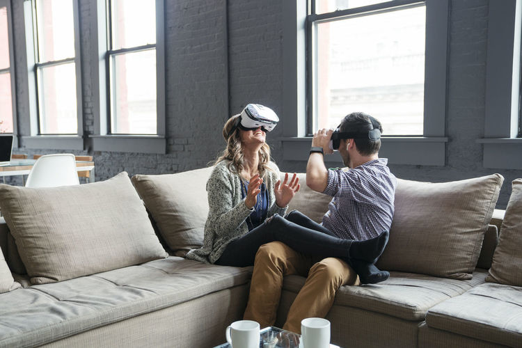 Happy business people wearing virtual reality simulator while sitting on sofa in office
