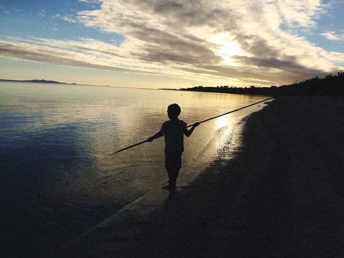 Silhouette boy fishing in sea against cloudy sky