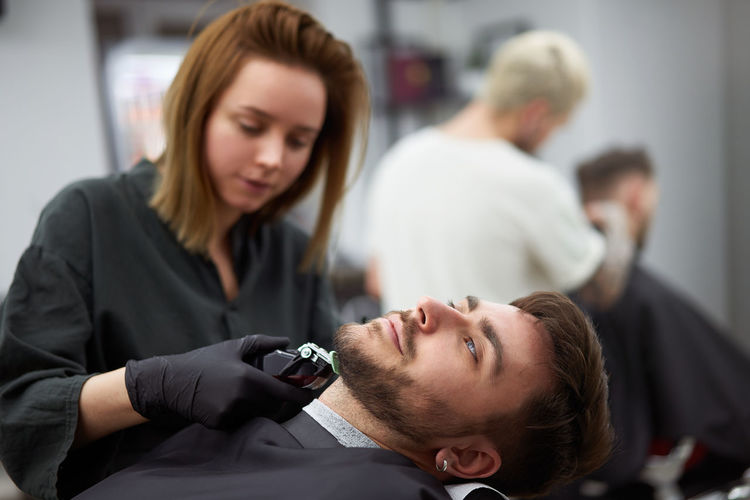 Portrait of young man who sitting in barbershop and woman hairdresser cut hair