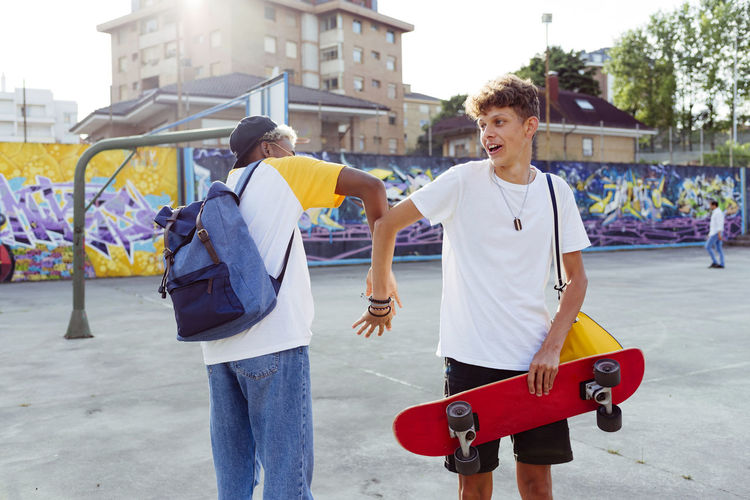 Two teenage boys with skateboard and backpack shaking hands and laughing on the street