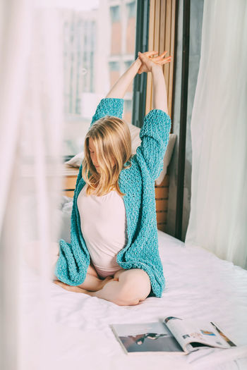A young girl in pyjamas and a blue knitted cardigan is sitting cross-legged on the bed, stretching.