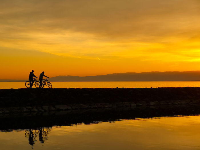 Silhouette people riding by lake against sky during sunset