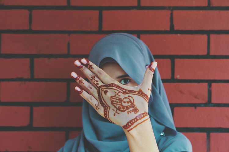 Portrait of young woman with henna tattoo hiding face against wall