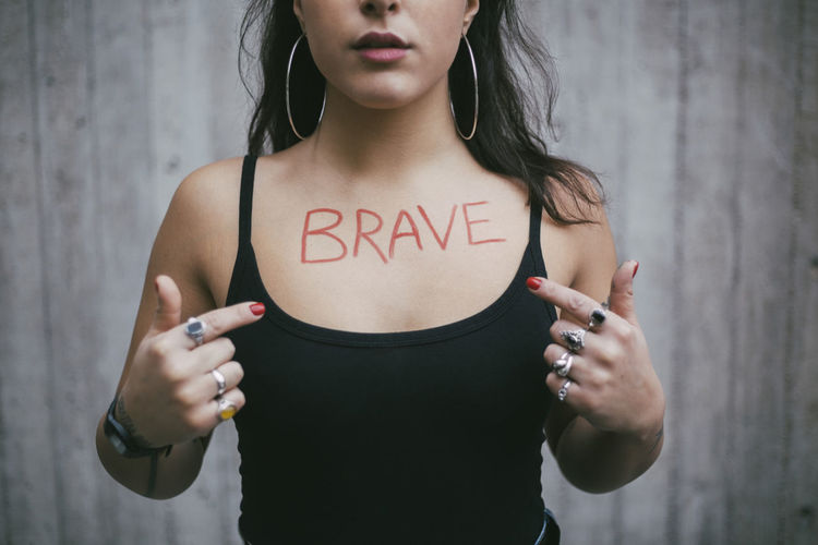 Midsection of female protestor pointing on brave text written over chest against wall