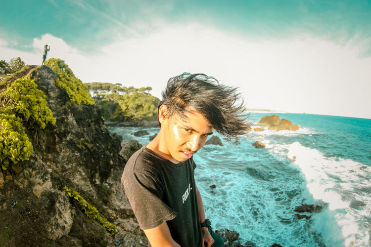 Portrait of young man standing by cliff against sea
