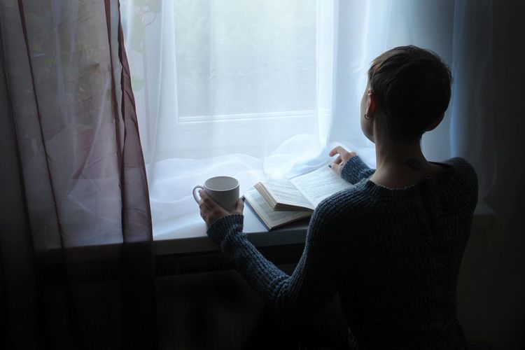 High angle view of woman holding coffee cup while reading book on window sill