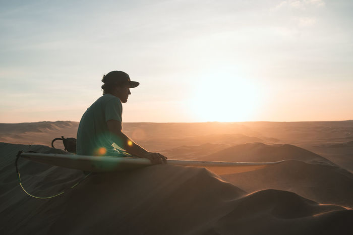 Man with sandboard sitting at desert during sunset